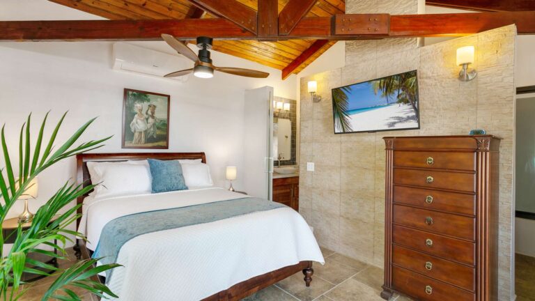 Bedroom with a wooden sleigh bed, vaulted timber-beam ceiling, ceiling fan, wall-mounted TV and a tall wooden dresser against a stone accent wall.