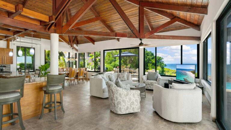 Open-plan great room with a vaulted timber cathedral ceiling, white sofas and patterned swivel chairs, a bar and walls of glass framing the sea.