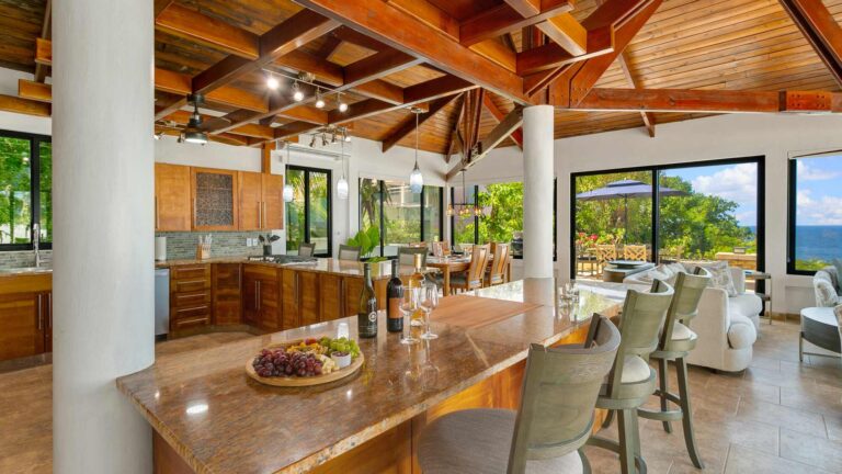 Granite-topped kitchen island with a cheese and wine spread and bar stools, open to the living area with ocean views beyond.