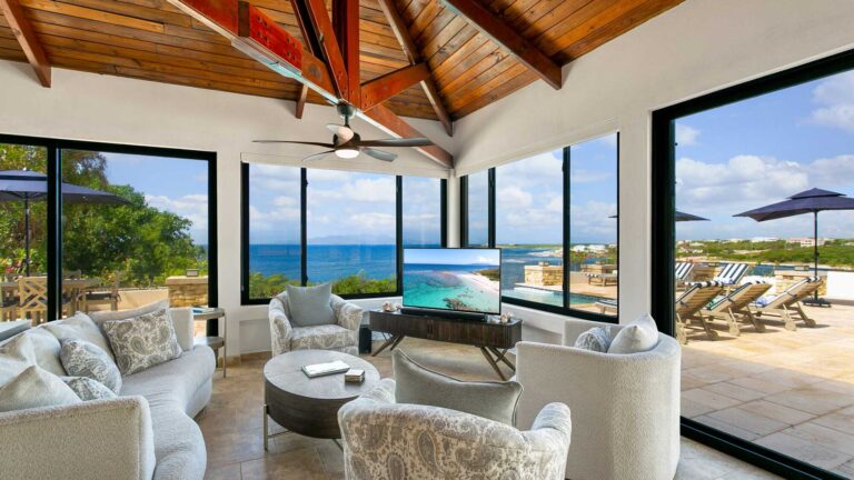 Sitting area with white sofas and patterned armchairs around a round table, with corner glass walls framing the sea and a pool terrace outside.