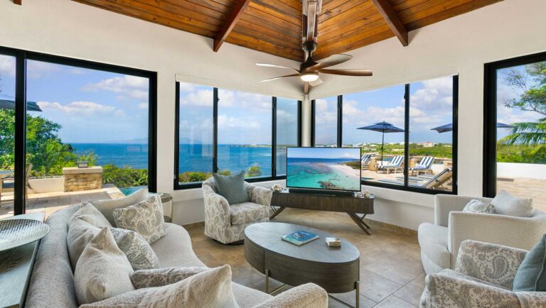 Lounge with cushioned sofas and swivel chairs, a wood-beamed ceiling and walls of glass overlooking the sea and a pool terrace with loungers.