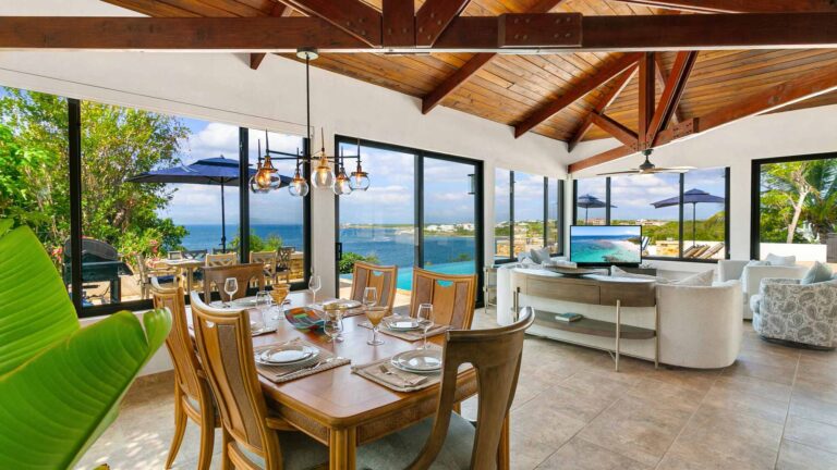 Wooden dining table set for six beneath glass pendant lights, open to the living area with an infinity pool and ocean beyond.