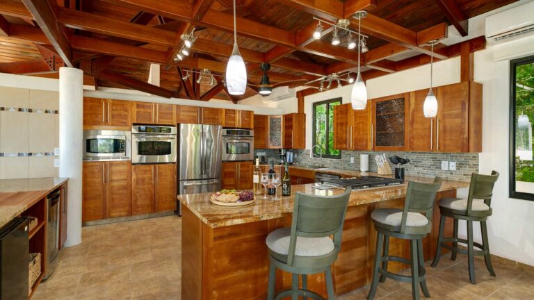 Kitchen with warm wood cabinetry, stainless steel double wall ovens and refrigerator, a granite island with bar stools and a vaulted timber ceiling.