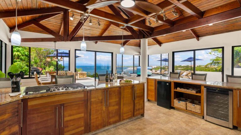 Kitchen island with a gas cooktop and granite counter, a wine fridge and beverage station, framed by glass walls overlooking the sea and pool terrace.