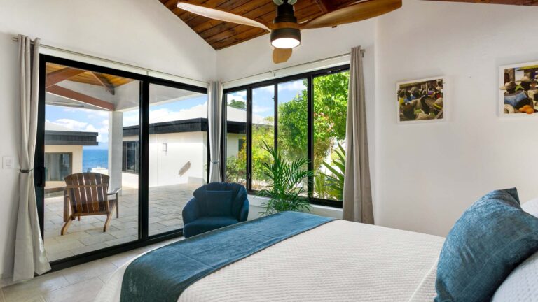 Bedroom with a vaulted wood ceiling and sliding glass doors opening to a terrace with sea views, and a blue accent chair beside the bed.
