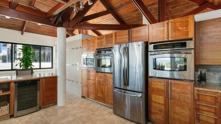 Kitchen with wood cabinetry, stainless steel double wall ovens, a French-door refrigerator and a built-in wine fridge under a vaulted timber ceiling.