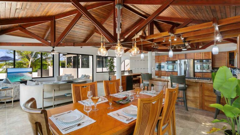 Wooden dining table set for six with glass pendant lights, open to the kitchen and a lounge with a TV beneath a vaulted timber ceiling.