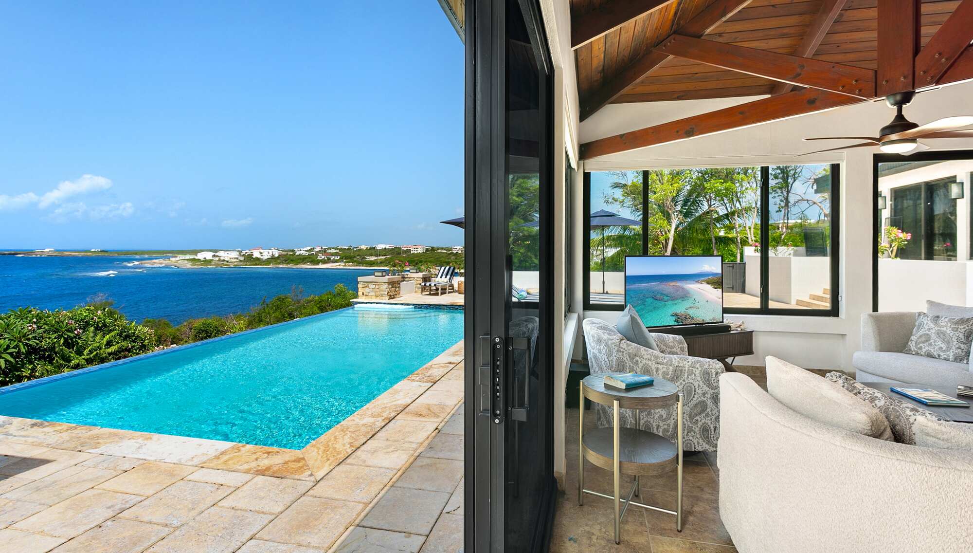 Covered terrace with cushioned seating opening to an infinity pool that overlooks the sea and coastline, with a timber ceiling and ceiling fan.