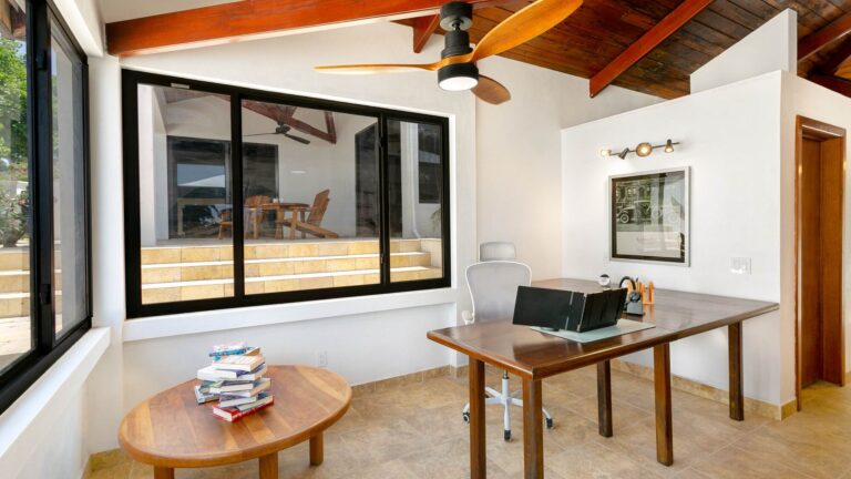 Home office with a wooden desk and laptop, a white ergonomic chair, framed vintage-car artwork and an interior window onto the dining terrace.