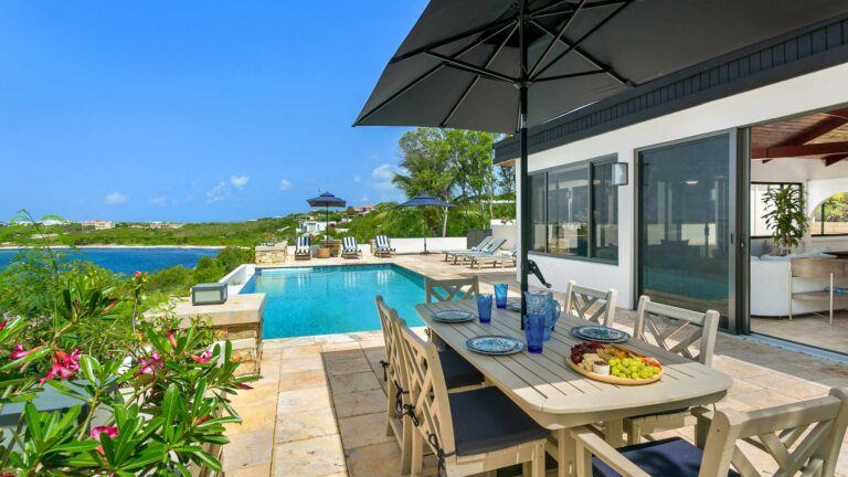Outdoor dining table set on a travertine terrace under a large umbrella, beside an infinity pool with sea views and flowering plants.
