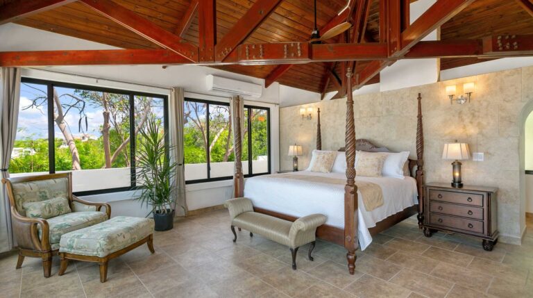 Master bedroom with a carved mahogany four-poster bed, vaulted timber ceiling, large garden windows, an armchair with ottoman and a stone accent wall.