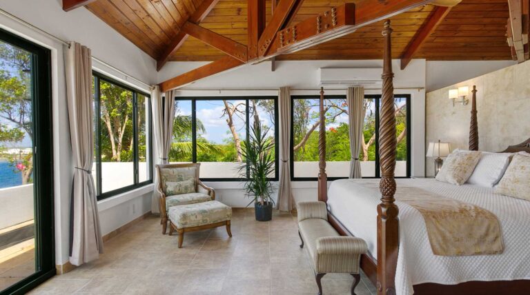 Bedroom with a mahogany four-poster bed, corner glass walls framing the sea, an upholstered armchair with ottoman and a vaulted timber ceiling.