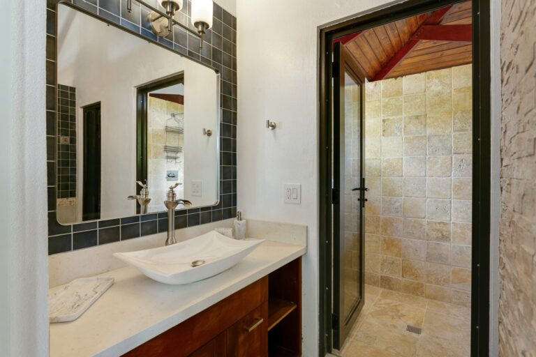 Bathroom with a white vessel sink on a wood vanity, a blue-tiled mirror surround and a glass-door travertine walk-in shower.