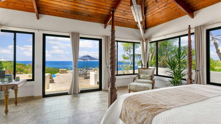 Bedroom with a four-poster bed and sliding glass doors opening to a terrace with an umbrella, pool and sea views, plus an upholstered armchair.