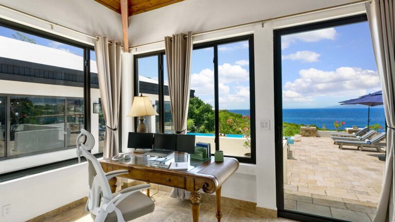Study with a wooden desk, dual monitors and an ergonomic chair, framed by glass walls overlooking the pool terrace and sea.