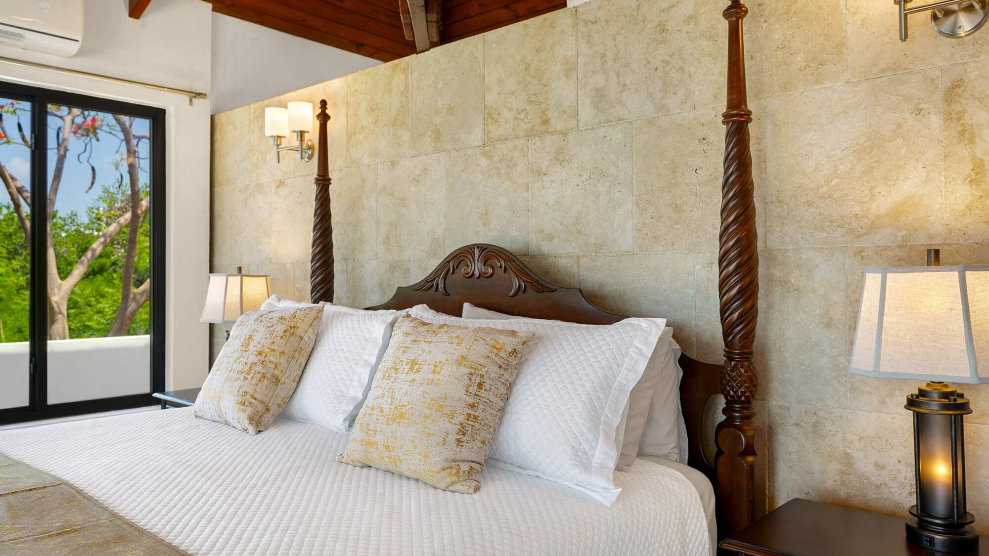 Close view of a carved mahogany four-poster bed with gold and white pillows against a stone feature wall, with lantern-style bedside lamps and a garden door.