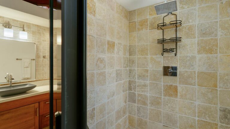 Travertine-tiled walk-in shower with a rainfall showerhead, beside a wood vanity with a vessel sink and mirror.