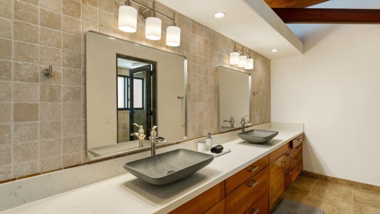 Bathroom with a long double vanity holding two stone vessel sinks, a travertine backsplash, twin mirrors and cylinder pendant lights.