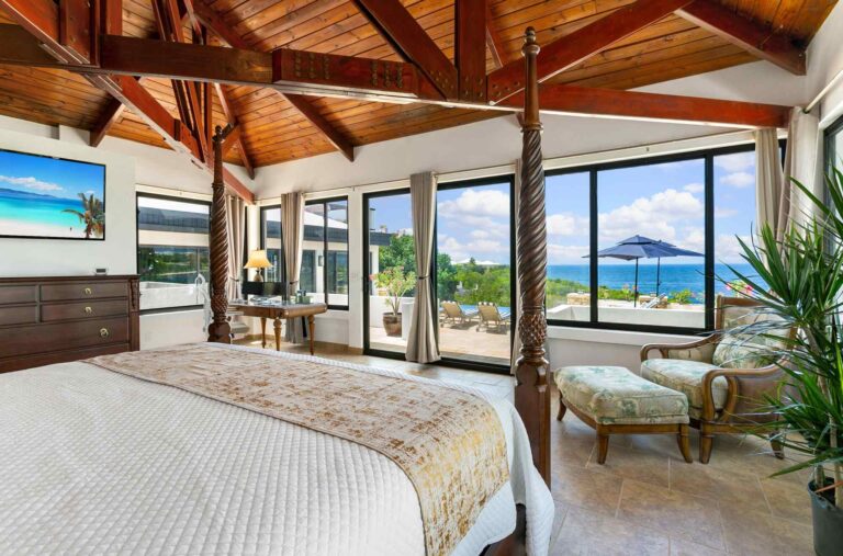 Master bedroom with a four-poster bed, vaulted timber ceiling, wall-mounted TV and walls of glass opening to a terrace with sea views and a pool umbrella.