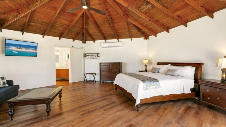 Spacious bedroom with a vaulted timber ceiling and wood floor, a sleigh bed, dresser, wall-mounted TV, a sofa and coffee table and an ensuite behind a barn door.