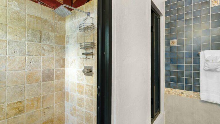 Walk-in shower with travertine stone walls, a rainfall showerhead, a wire caddy and a blue mosaic tile accent with white towels.
