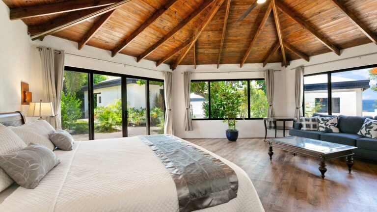 Bedroom with a vaulted timber ceiling and wood floor, a bed with a silver runner, sliding glass doors to a garden, and a blue sofa and coffee table.