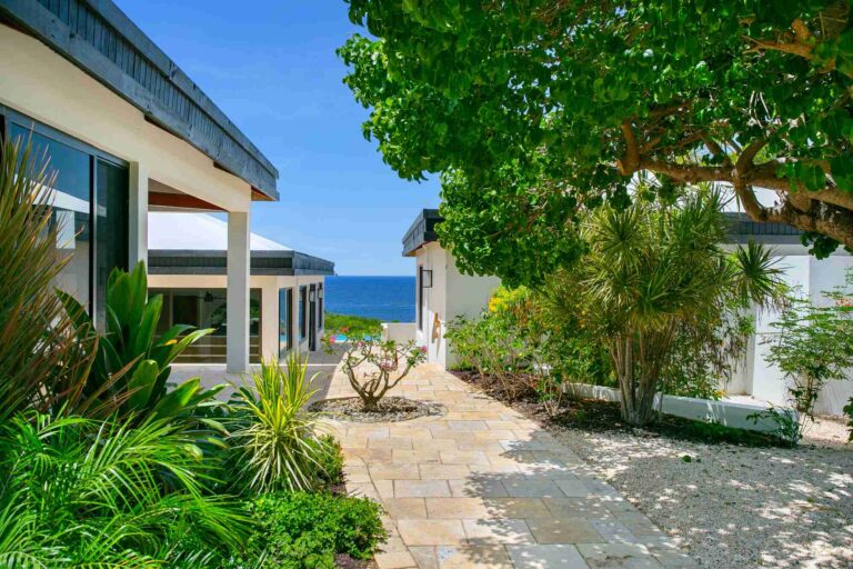 Exterior of the villa with a colonnaded covered terrace and a travertine path through tropical landscaping, overlooking the sea.