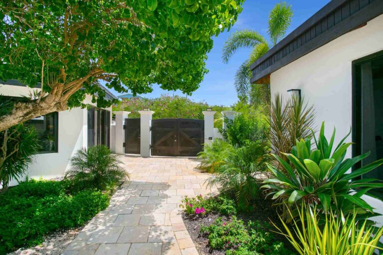 Entrance courtyard with a dark timber double gate, a travertine path, flowering shrubs and tropical plants between the villa wings.