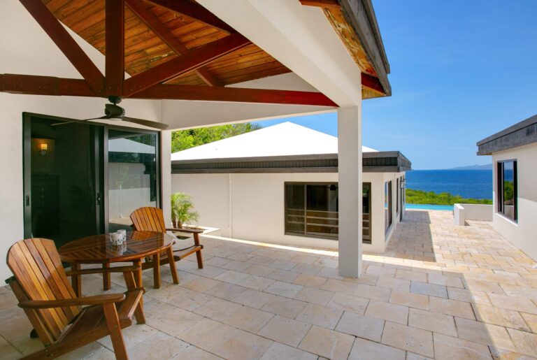 Covered travertine terrace with Adirondack chairs and a wood table, columns framing the sea and a glimpse of the pool, beside the villa wing.