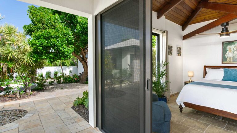 Bedroom with a sliding screen door opening to a travertine garden patio, with tropical plants outside and a bed with blue accents.