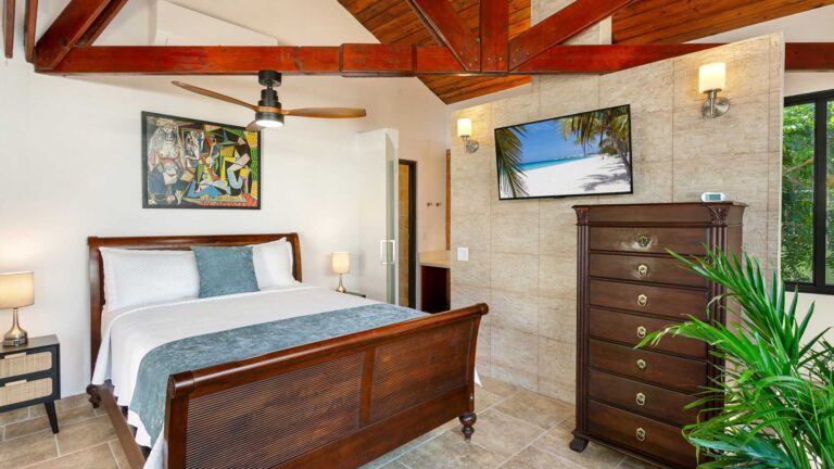 Bedroom with a wooden sleigh bed, vaulted timber ceiling, framed cubist artwork, a wall-mounted TV and a tall dresser by a stone wall.