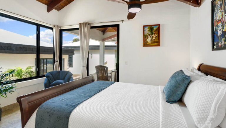 Bedroom with corner windows overlooking a courtyard and the sea, a wooden sleigh bed with a blue throw and a blue armchair.